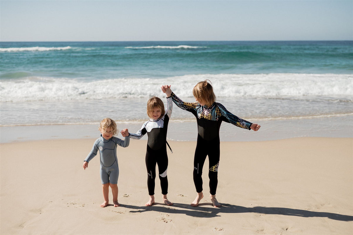 Three kids holding their hands up, in wetsuits.